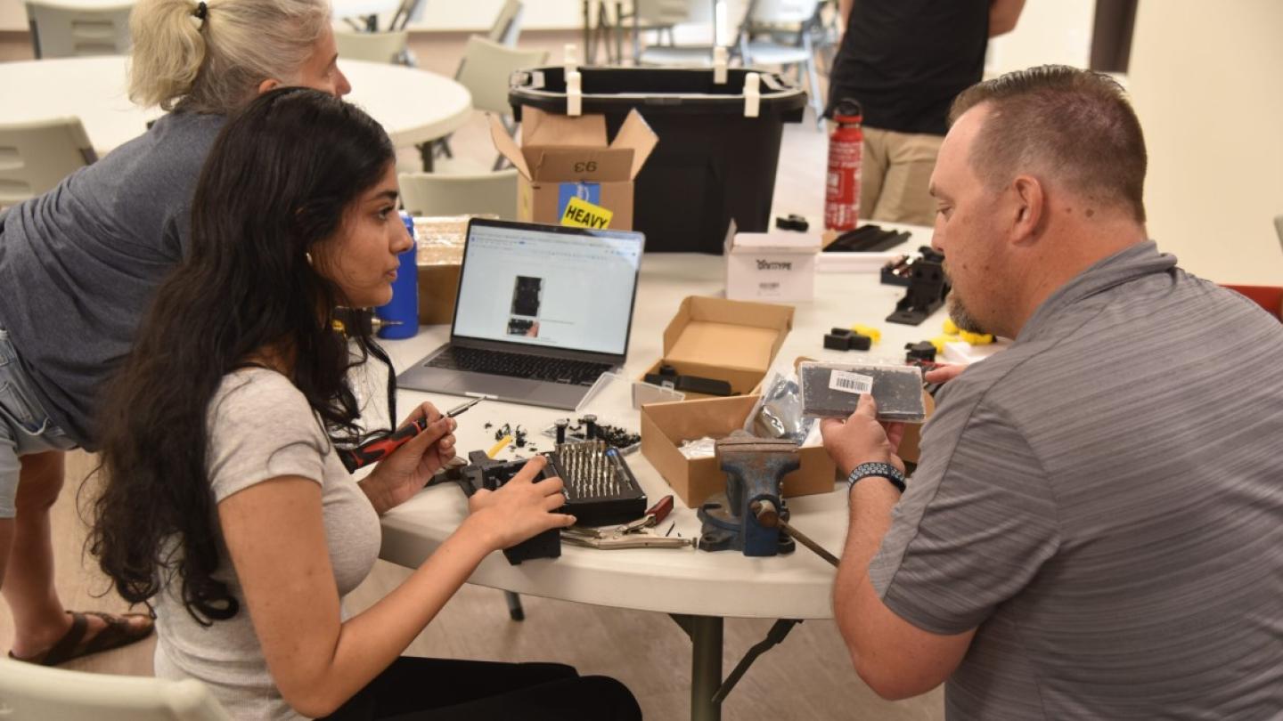 Two people sit at a table loaded with equipment, assembling a robot