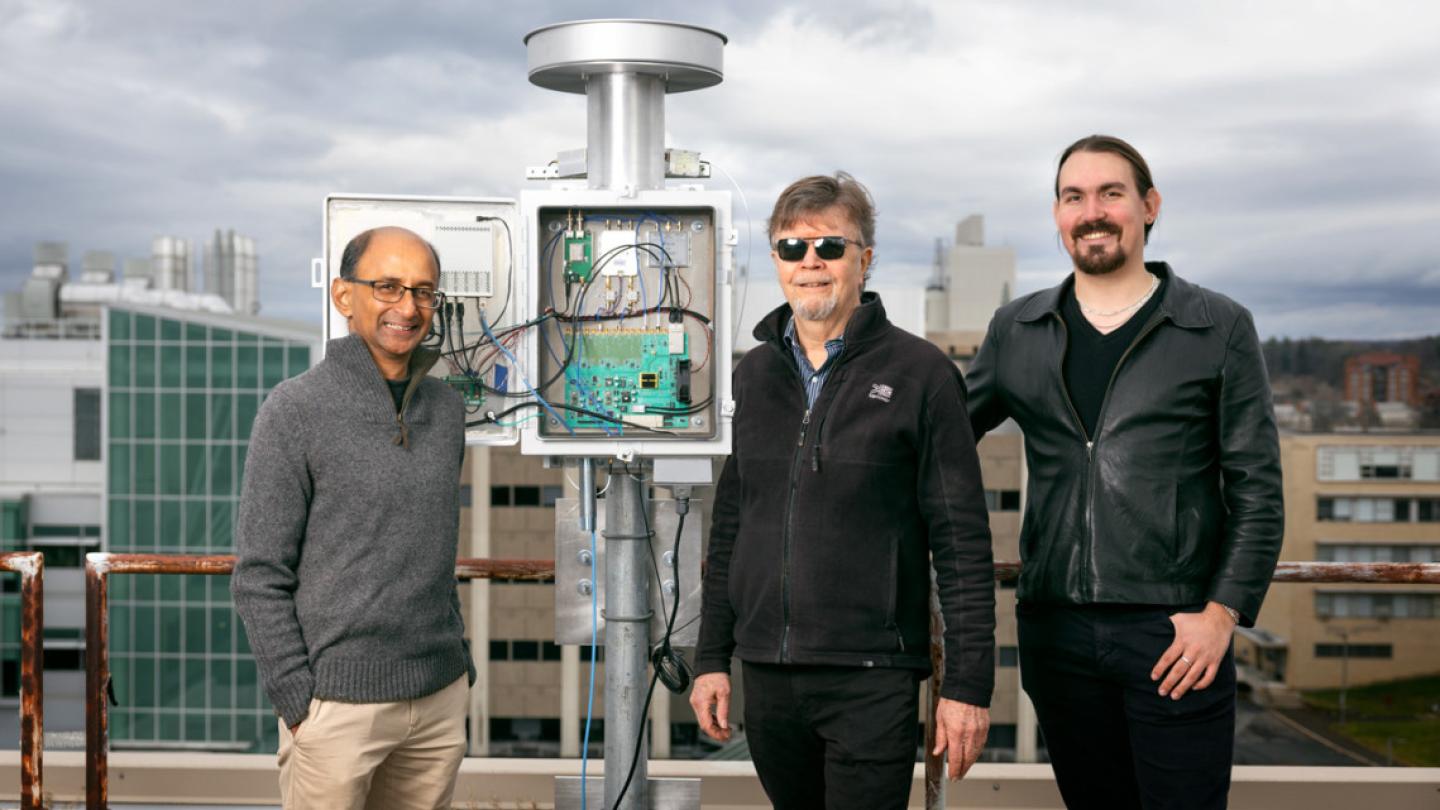 Three people stand next to a silver device, about a foot taller than the people, made of a post, a box with electronics inside, and a circular top