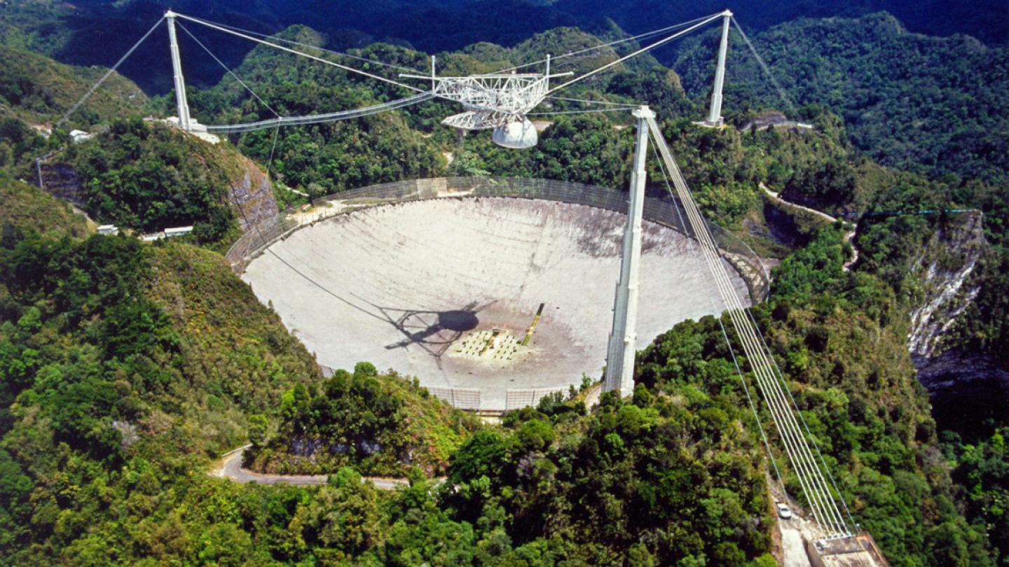 Large concrete dish surrounded by three poles and wires; a mechanism is suspended over the dish. the whole thing, a telescope, is surrounded by lush trees