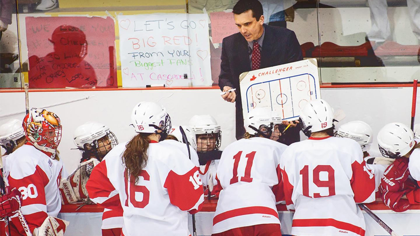 A coach with a white board in front of several hockey players in red and white jerseys