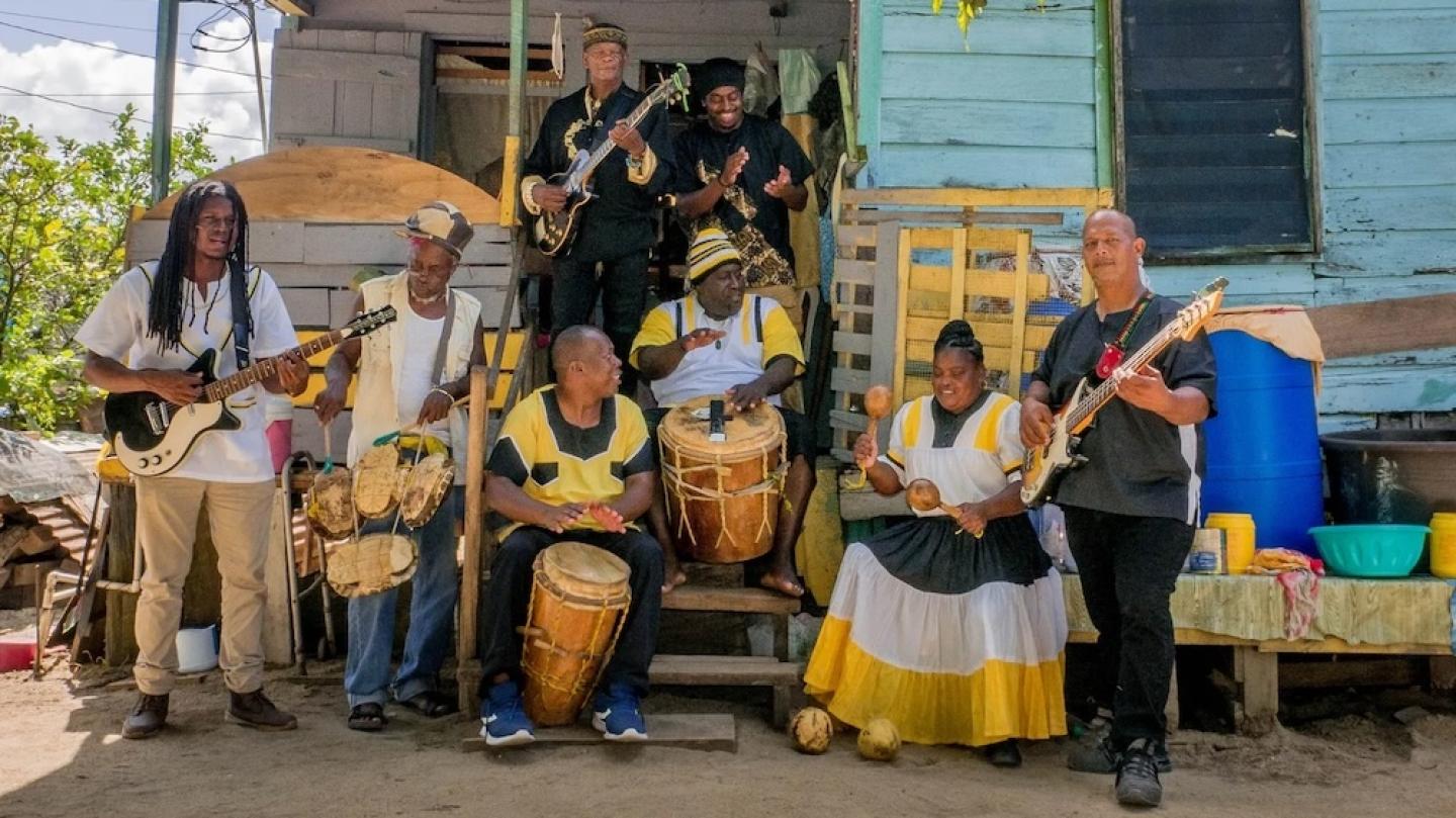 Several people standing around and sitting on porch steps in the shade, playing drums, guitars and maracas
