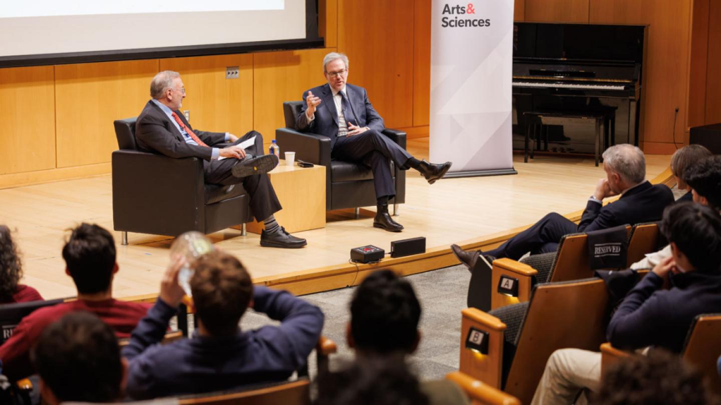 Two people seated in large chairs, conversing in front of an audience