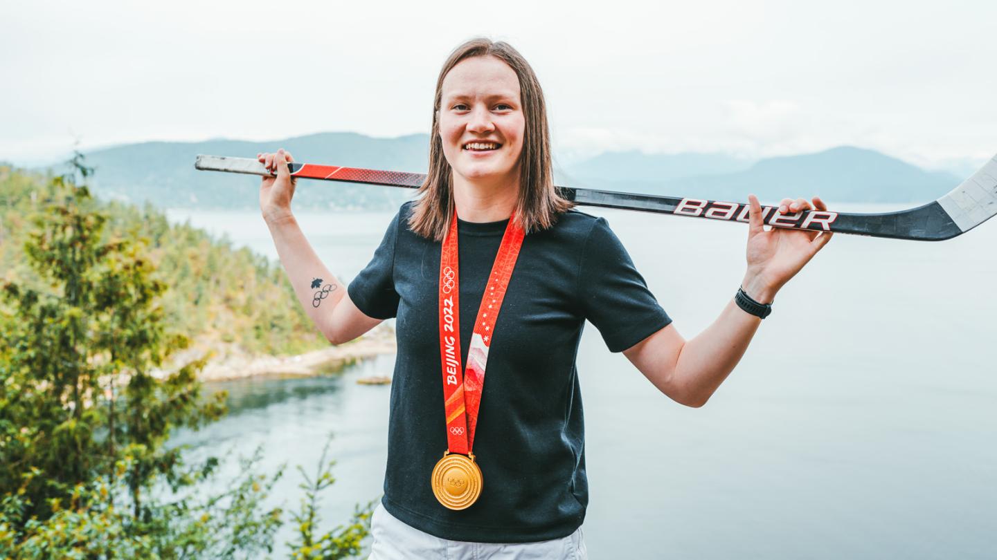 Person standing in front of a body of water, balancing a hockey stick across her back and wearing a gold medal on a red ribbon