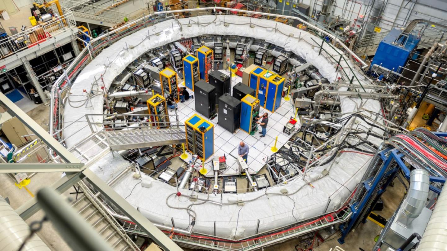 Very large, while ring – a physics experiment setup – dwarfs equipment in its center and people working in it 
