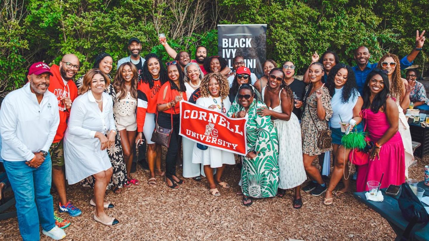 A few dozen people in summer fashions cluster together with a Cornell University banner